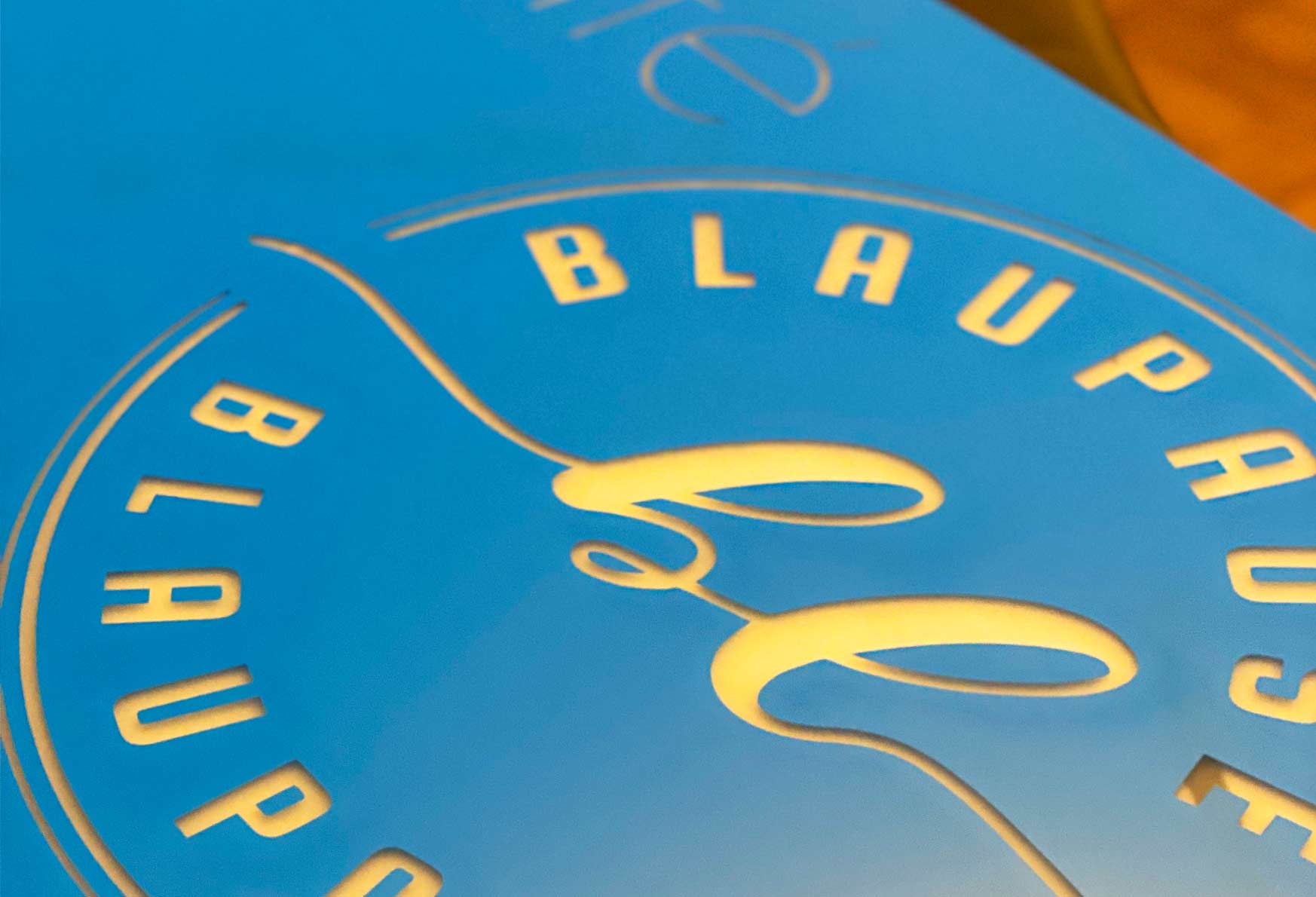 Nahaufnahme eines blauen, folierten Schildes, das in einem Kreis die Buchstaben 'Blaupause' und ein geschwungenes Logo erkennen lässt.