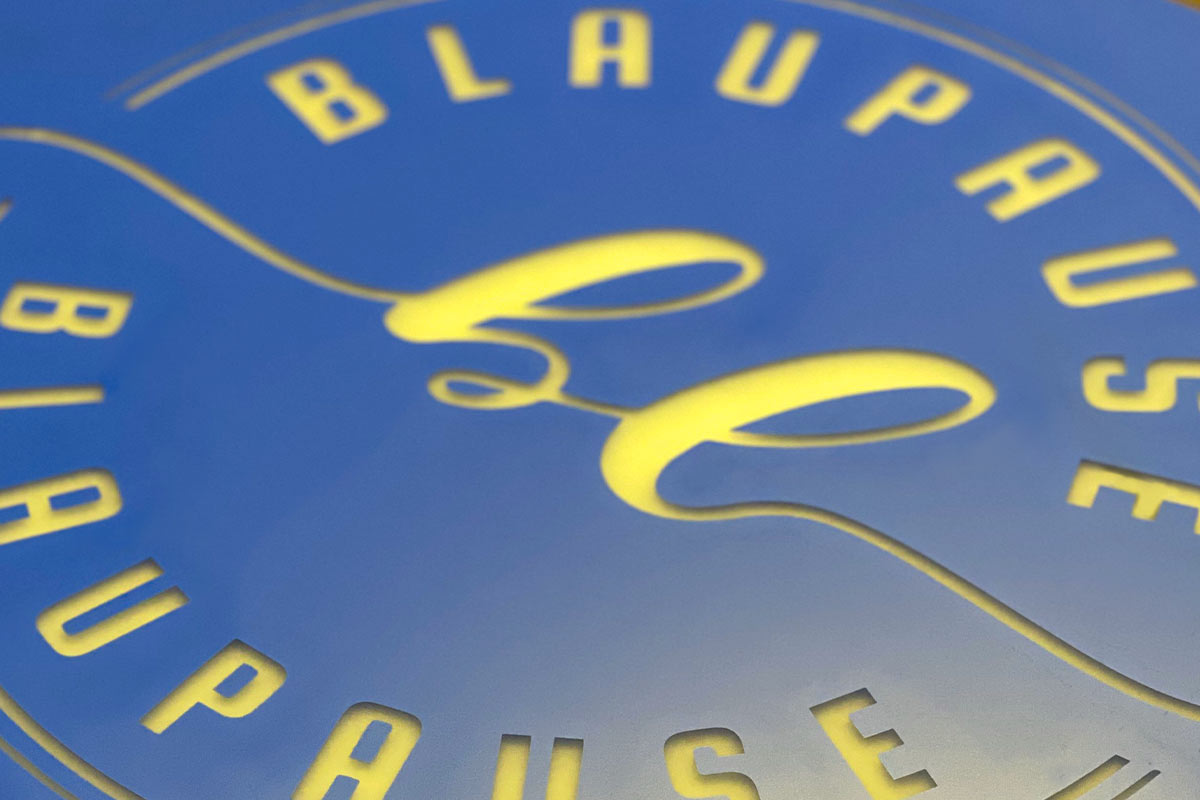 Nahaufnahme eines blauen, folierten Schildes, das in einem Kreis die Buchstaben 'Blaupause' und ein geschwungenes Logo erkennen lässt.